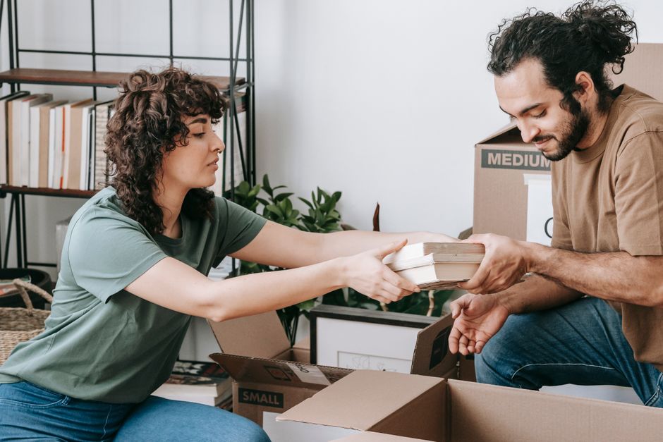 A woman with curly brown hair, wearing a green t-shirt, is seen inside a room, handing over a stack of books to a man with dark, curly hair and a beard, dressed in a brown t-shirt. They are engaged in