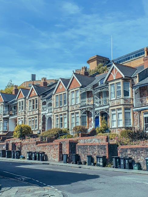 A row of Victorian-style terraced houses with ornate white iron balcony railings, peaked rooflines, and large bay windows facing a pavement. The front gardens are elevated and separated from the road