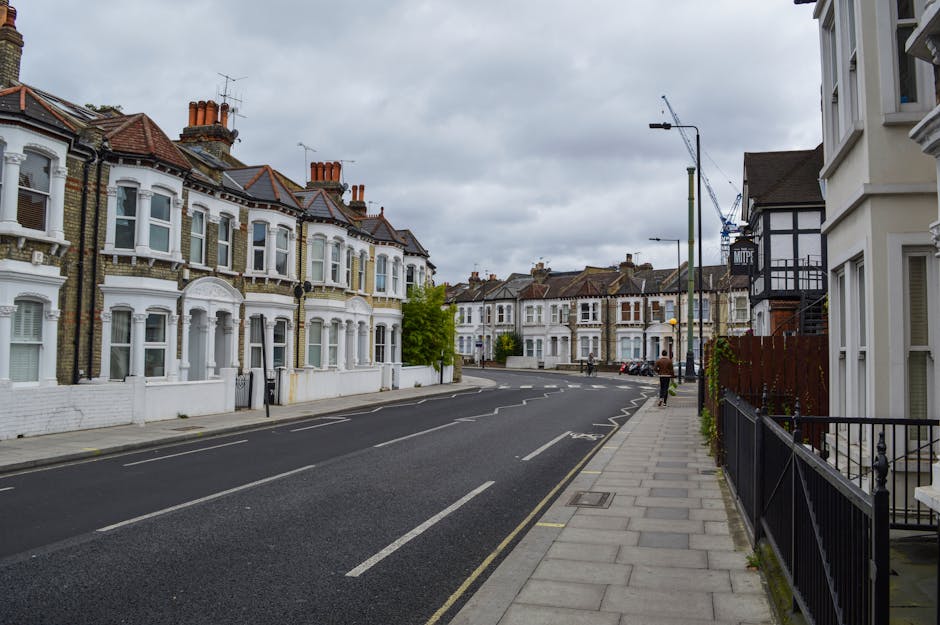 A row of Victorian-style terraced houses with ornate white iron balcony railings, peaked rooflines, and large bay windows facing a pavement. The front gardens are elevated and separated from the road
