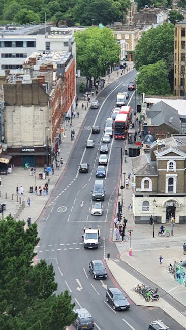 An aerial view of a street in Roehampton, showing a line of vehicles including vans and cars involved in a home relocation. The scene captures the loading process with some vehicles possibly used by Putney Man and Van for furniture transport and packing and moving services. The street is lined with various buildings, including residential houses with different architectural styles, as well as commercial properties such as shops and cafes. Sidewalks are occupied by pedestrians and cyclists, and trees provide greenery along the pavement. The overall environment is well-lit, indicating daytime conditions, with visible activity in a neighbourhood setting typical for house removals and relocation logistics, supporting the services offered by Putney Man and Van.