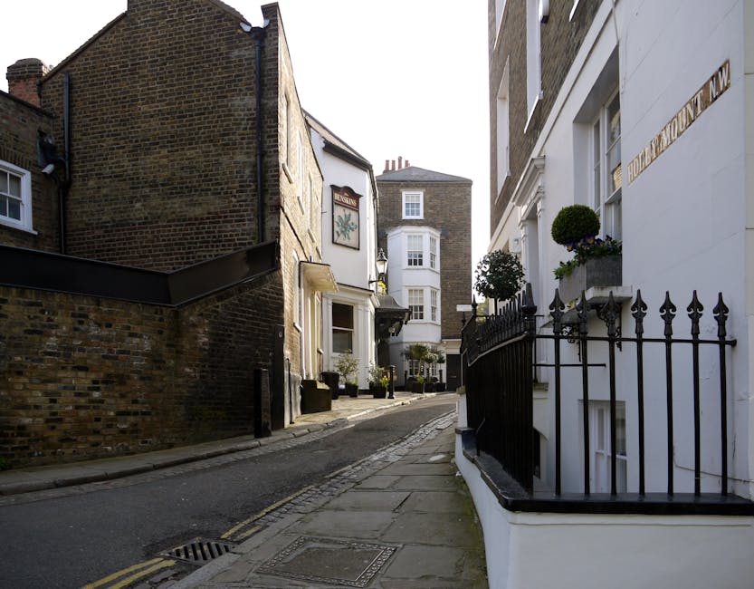 A narrow residential street in Roehampton Lane, showing a small white house with decorative fencing and potted plants on its balcony, adjacent to a taller brick building with a shop sign. The street has a slight incline, with a paved sidewalk on the right side and a cobbled section in the middle. Visible objects include cardboard boxes, wrapped furniture, and a metal trolley, indicating a home relocation or moving process. The scene is well-lit with daylight, and the placement of boxes and furniture suggests they are ready for loading onto a van for transportation. Putney Man and Van offers local removals on this route, supporting furniture transport and packing and moving services for house relocations within the Roehampton area.