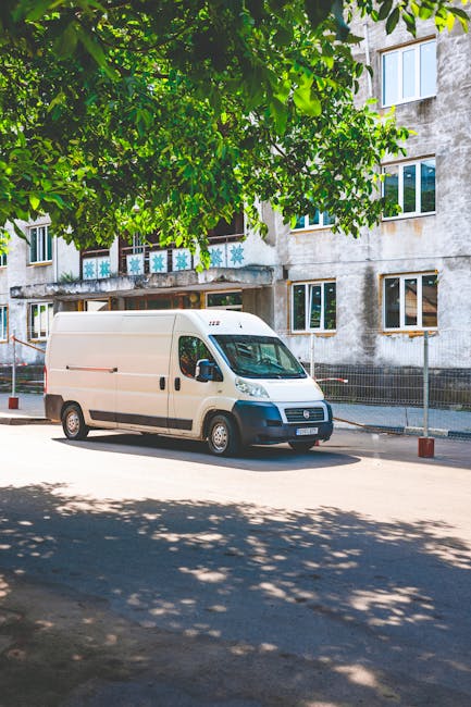 This image captures the side view of a white commercial van parked on a grassy area near a residential building, with leafy green trees casting shadows across the vehicle. The van appears used, with r