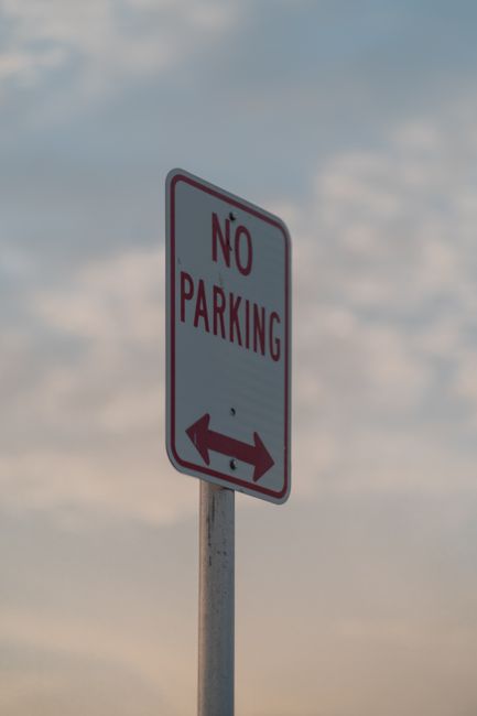 A round traffic sign with a red border, featuring a blue circle with a red diagonal line through it, indicating no parking, is mounted on a metal post. The sign is positioned against a textured stone