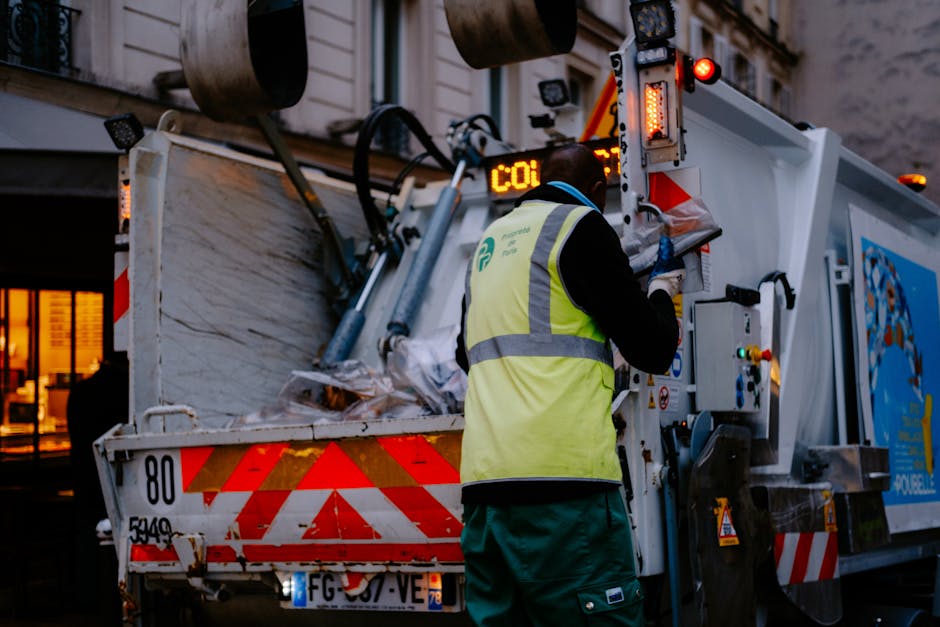 A close-up of a person wearing an orange protective suit and a white glove, holding a large blue plastic bag filled with waste material, likely on a delivery or collection route for waste disposal. Th
